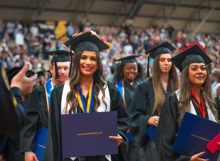毕业生们戴着帽子，穿着长袍站着. 最前面的毕业生拿着文凭微笑着.
